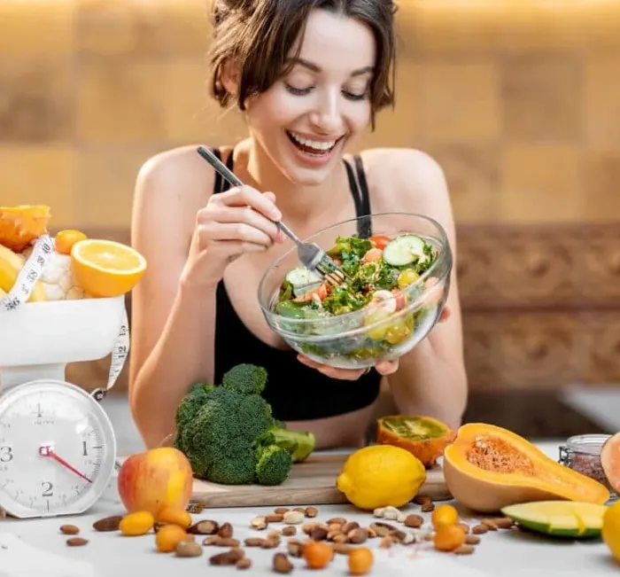 A smiling woman eating a large bowl of salad with fruit and vegetables